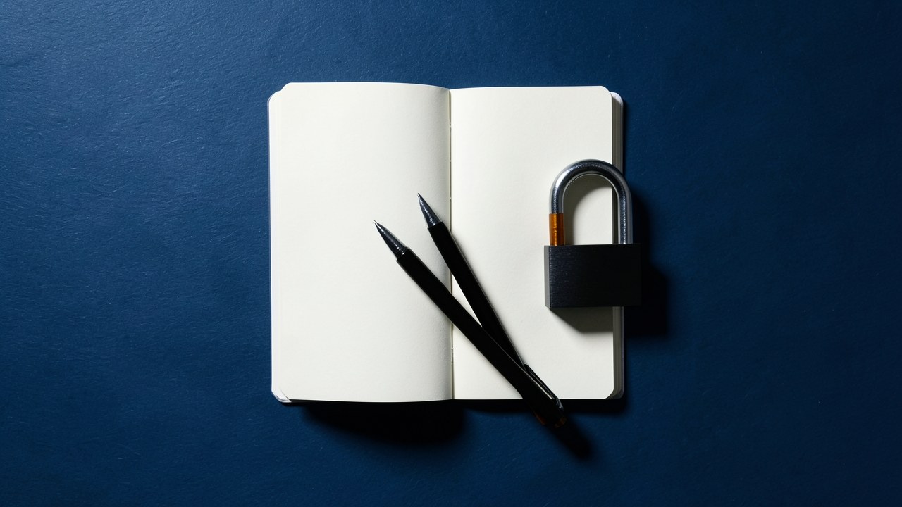 A monochrome flat lay of a black pen, a small notebook, and a padlock on a slate-blue surface, representing loan types and security.